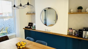Een moderne keuken met een houten eettafel, blauwe stoelen en een schaal met citroenen. Er zijn hanglampen, een ronde spiegel en een koffieapparaat op het aanrecht.