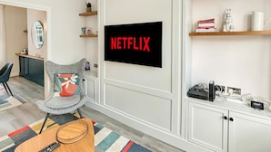 A living room with a large TV displaying "NETFLIX", a grey armchair, a small table with a remote, and a colorful striped rug.