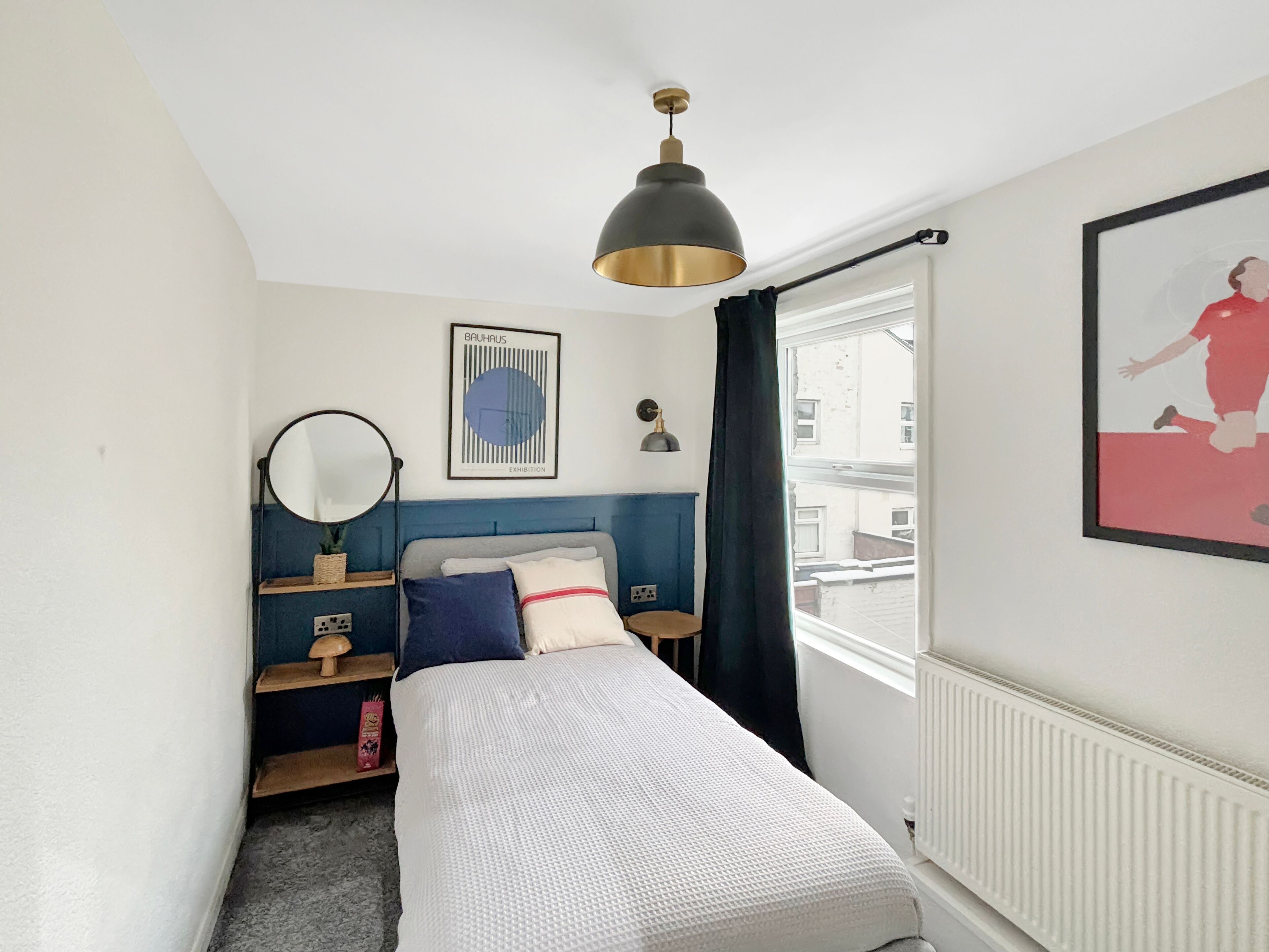 Une chambre à coucher comprenant un lit, une table de chevet, un miroir, un tableau encadré accroché au mur et une fenêtre avec des rideaux.