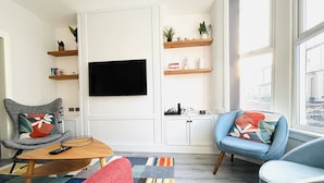 Een moderne woonkamer met een flatscreen televisie aan de muur, een houten salontafel, twee fauteuils en een kleurrijk vloerkleed.