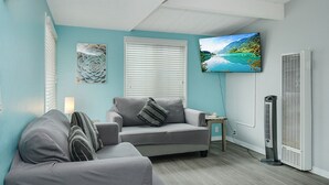 A living room with a grey sofa, a small table, a TV mounted on the wall, and a standing heater.