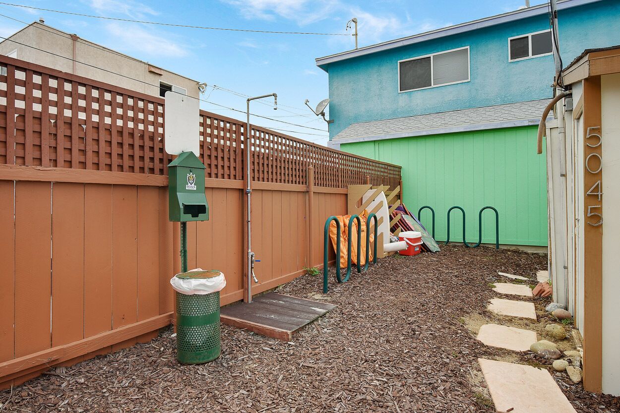 A fenced backyard with a green trash bin, a wooden step leading to a door, and a blue house in the background.