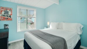 A bedroom with a large bed, a window with blinds, a nightstand with a lamp, and a poster on the wall.