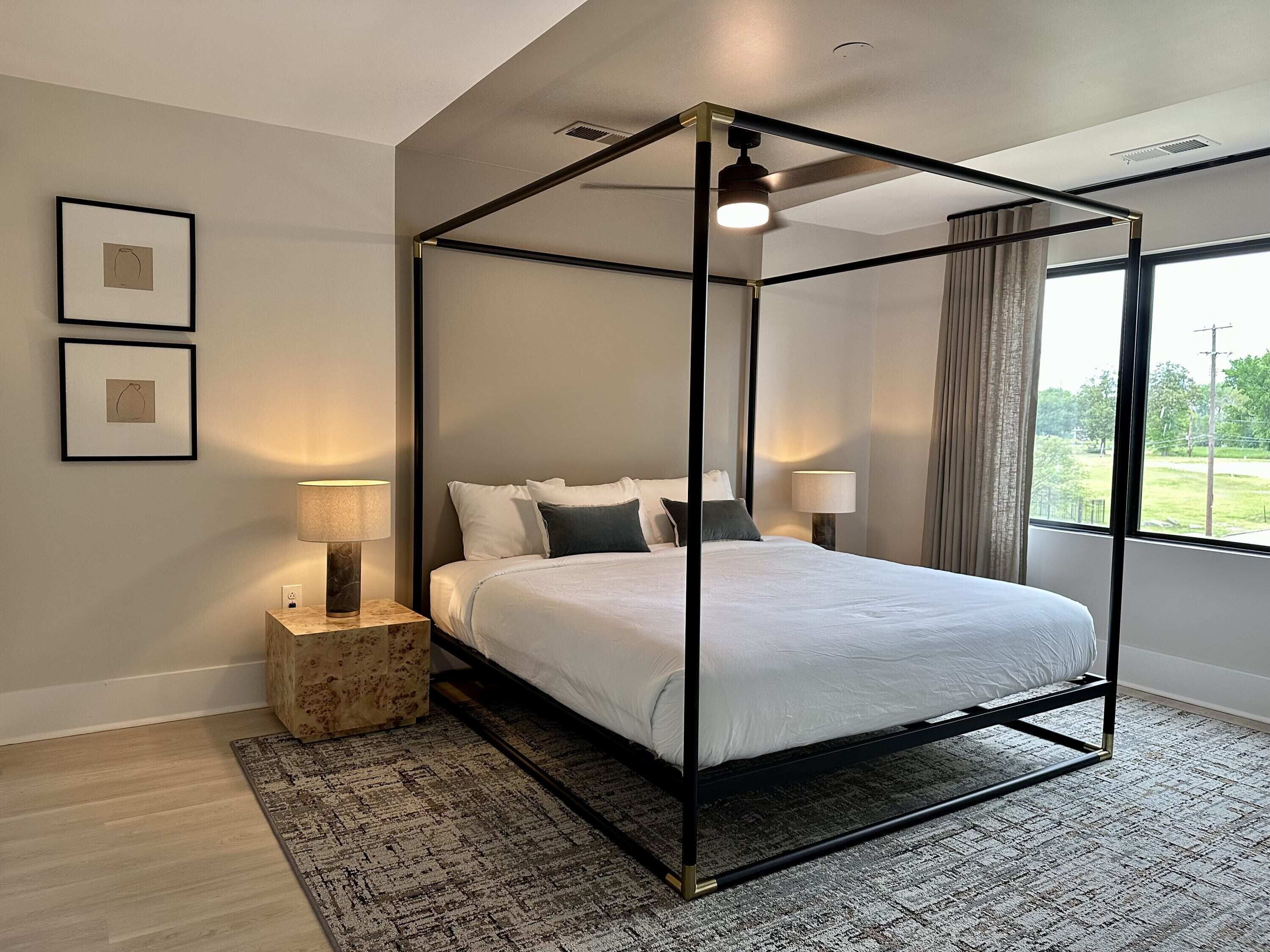 Un dormitorio moderno con cama con dosel, dos lámparas de noche, una mesita de madera pequeña, una alfombra estampada y cuadros en la pared.