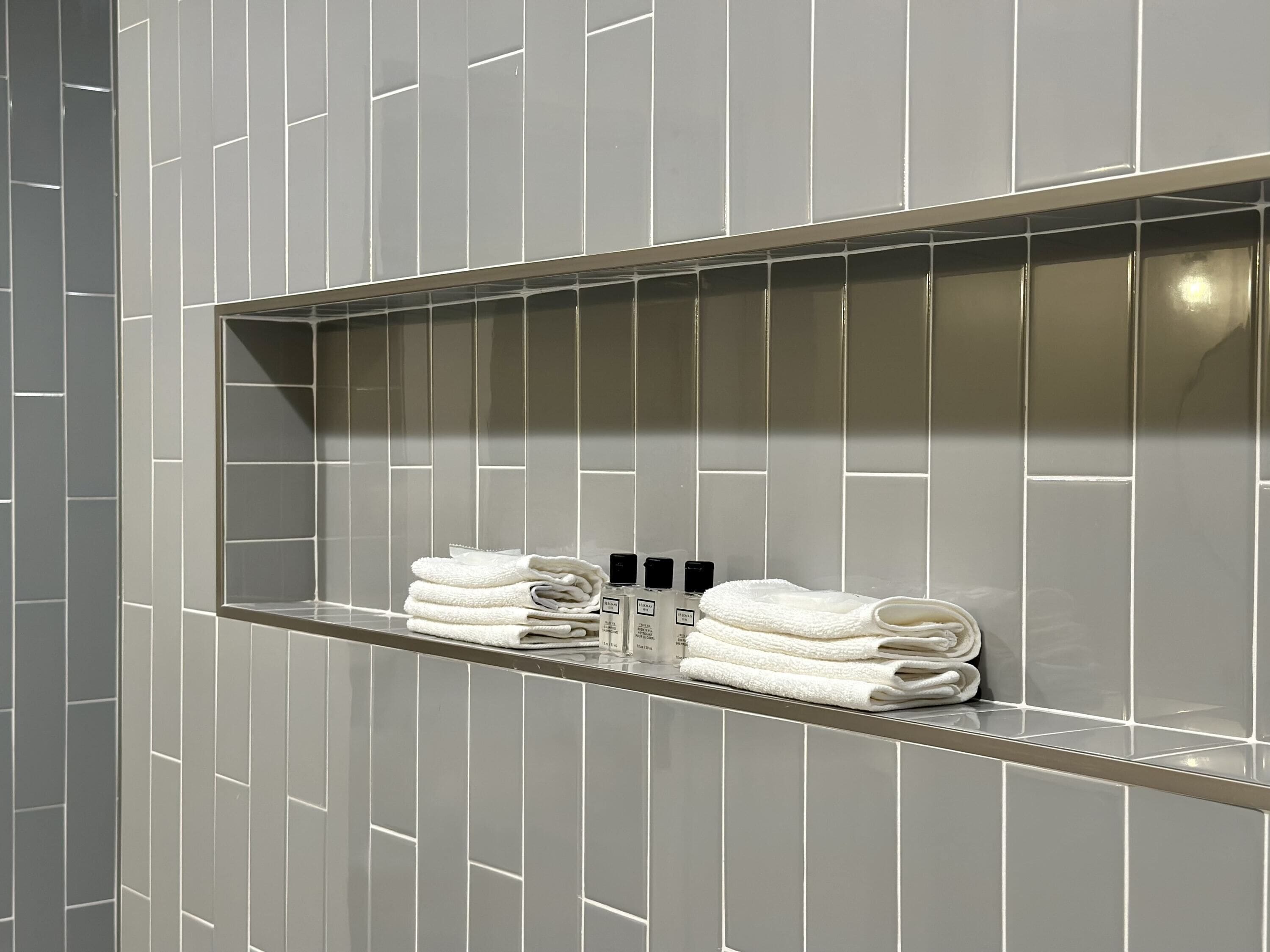 A bathroom with a tiled wall and a shelf holding folded towels and toiletries.