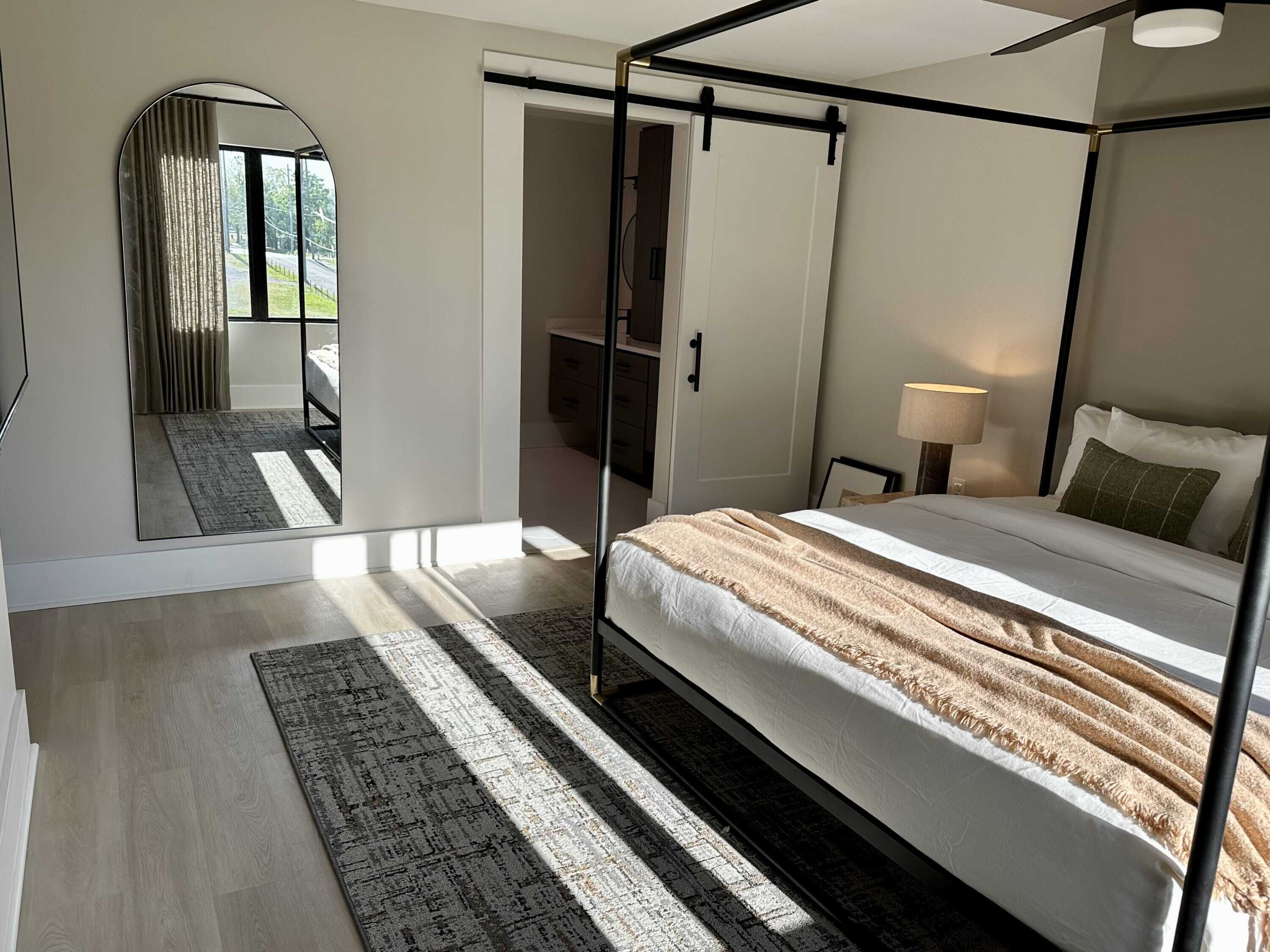 A four-poster bed with a white and beige striped blanket, a bedside lamp, and a view of a garden through a large window.
