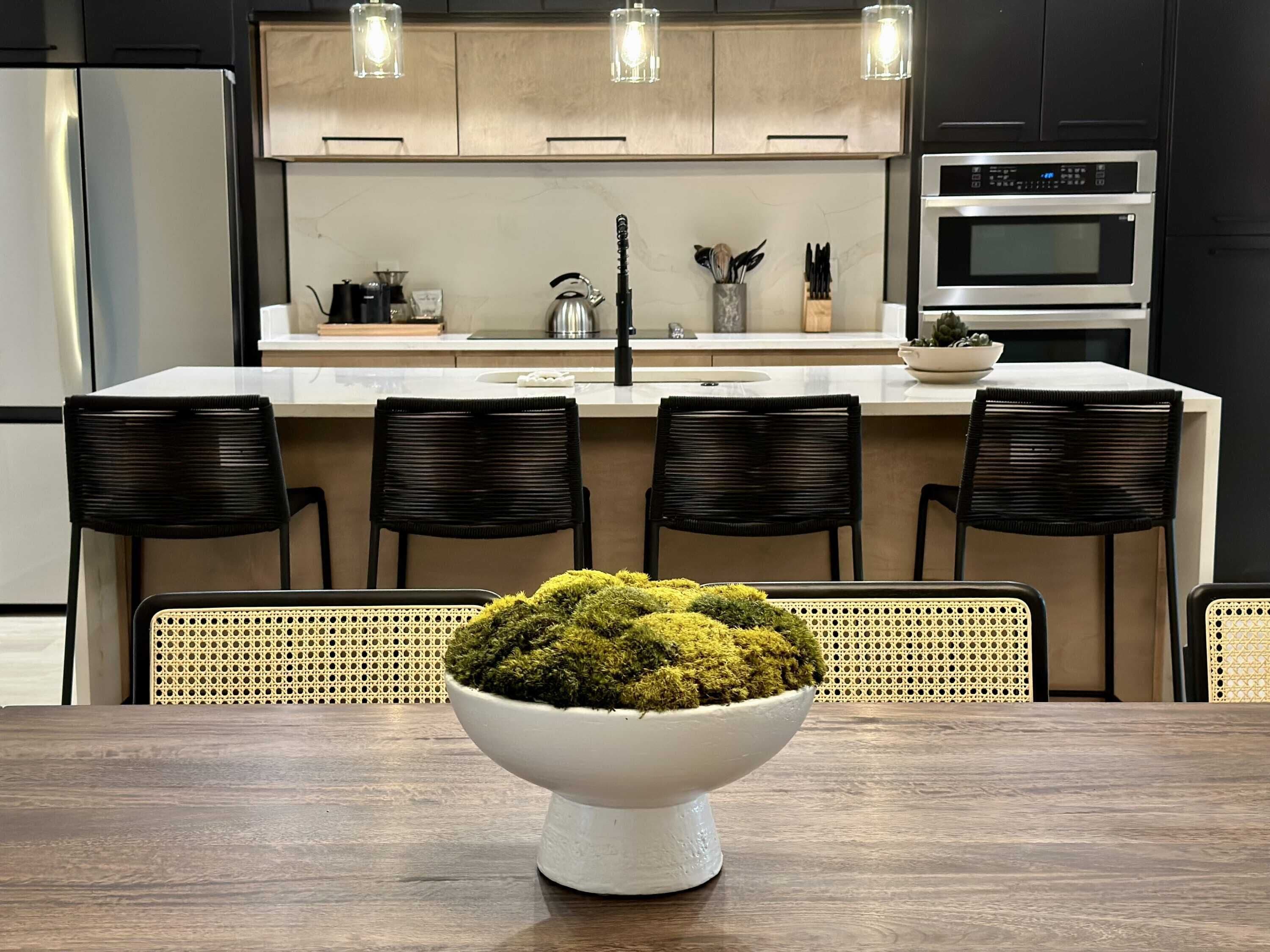 A modern kitchen with a central island, black chairs, and a white bowl with moss.