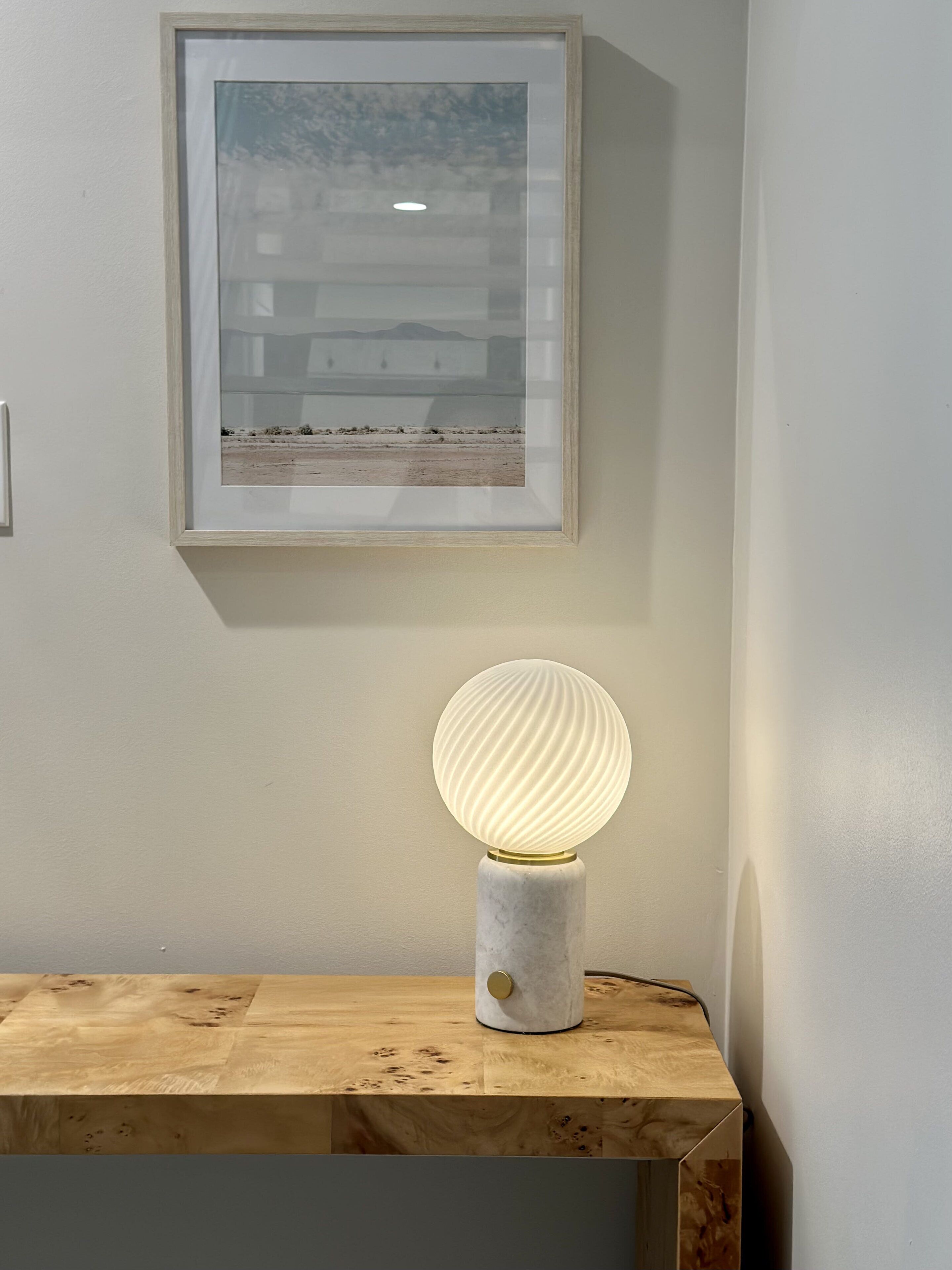 Un cuadro en la pared, una lámpara de mesa con base de globo texturizado y una mesa de madera.