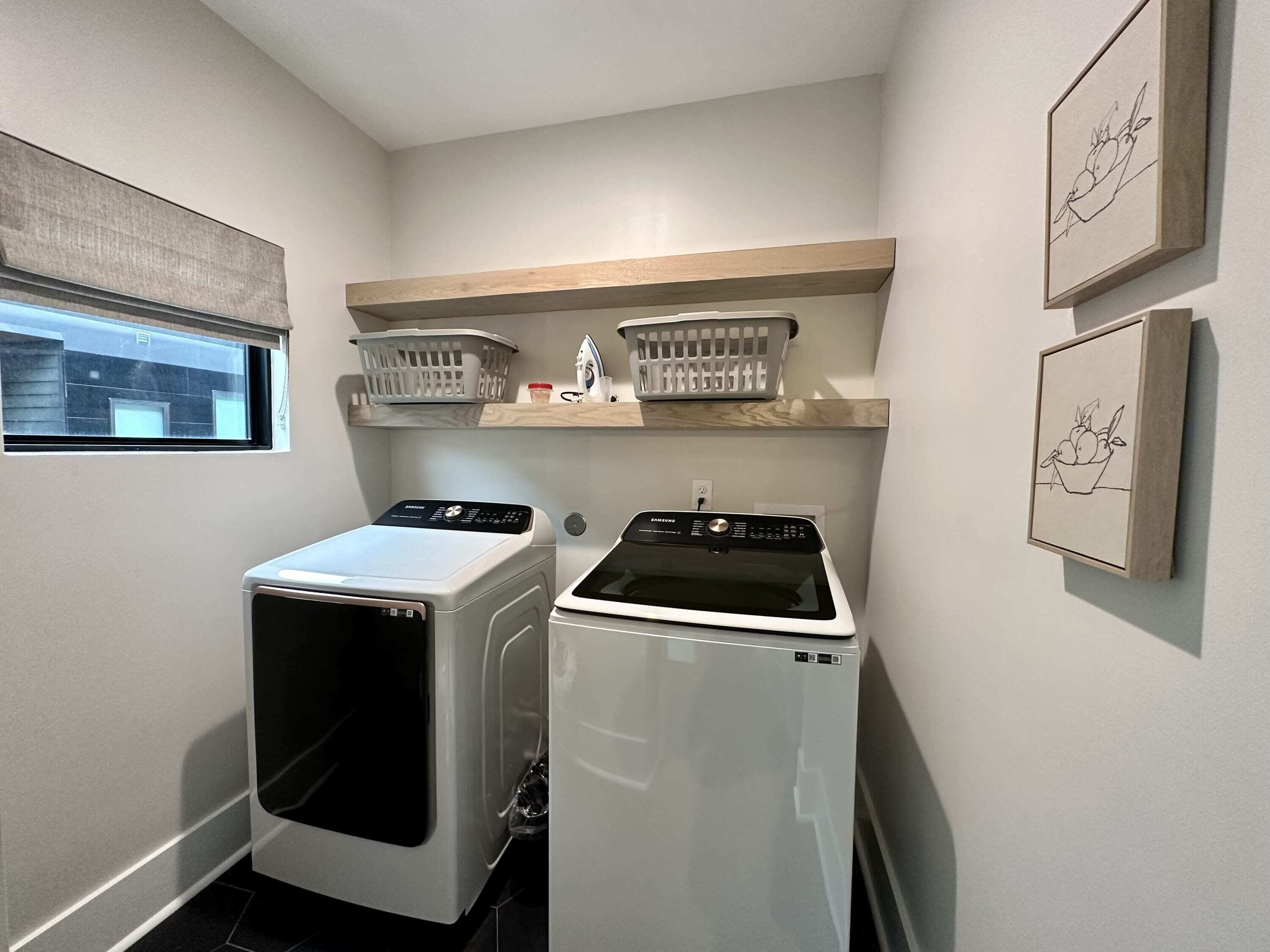 A laundry room with two front-loading washing machines, a window, and a shelf with baskets and a small decorative item.