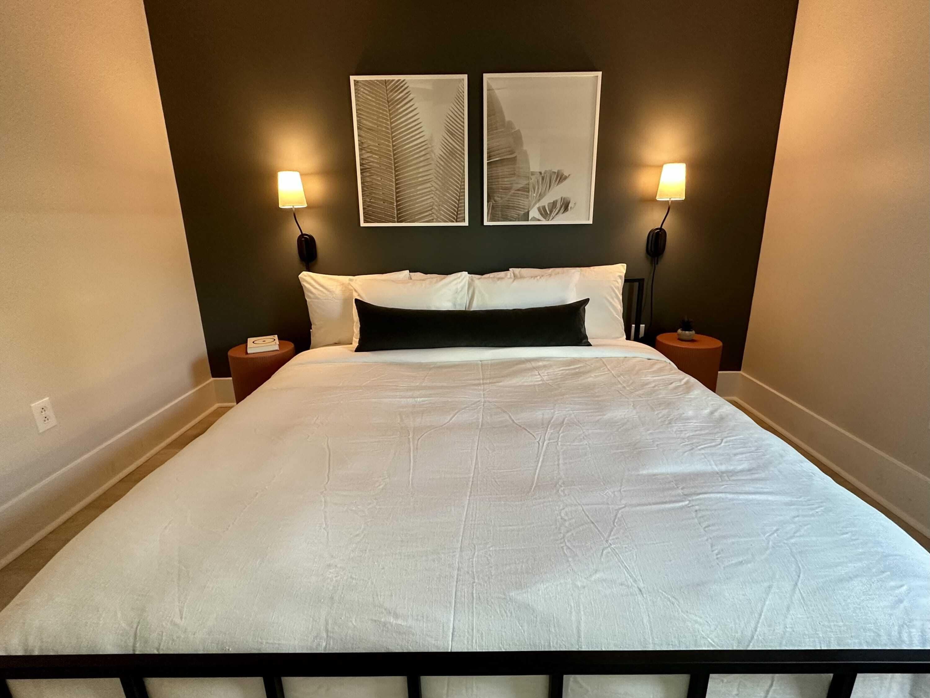 A neatly made bed with white linens, two wall-mounted lamps, and two framed leaf artworks on the wall.