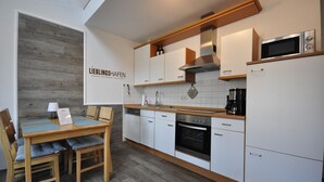 Een moderne keuken met witte kasten, een inbouwoven, een magnetron en een koffiezetapparaat. Er is een eethoek met een houten tafel en stoelen.