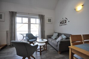 Ein modernes Wohnzimmer mit einem grauen Sofa, zwei Sesseln, einem kleinen runden Tisch und einer Wandleuchte.