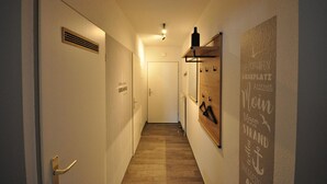 Een smalle gang met een houten vloer, een ventilatierooster aan de linkerwand en een deur aan het einde.