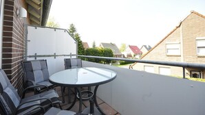 Een balkon met een glazen tafel en stoelen, een witte muur en een metalen hekwerk.