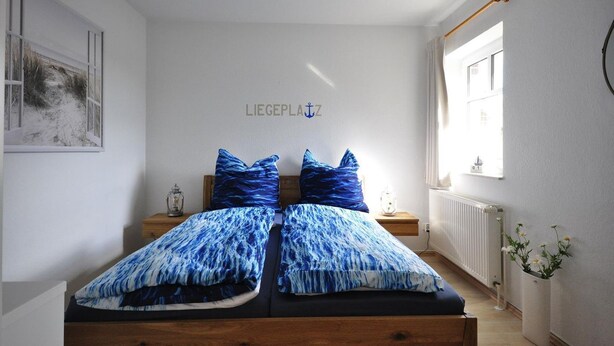 Een slaapkamer met een bed, twee nachtkastjes, een wandlamp, een raam met gordijnen, een radiator en een bordje aan de muur.