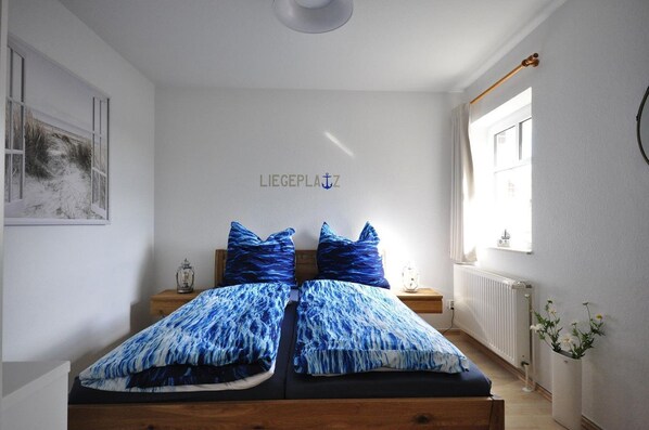 A bedroom with a bed, two nightstands, a wall-mounted light, a window with curtains, a radiator, and a wall-mounted sign.
