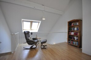 Une pièce spacieuse avec un plafond en pente, une lucarne, un fauteuil à bascule, une petite table et une bibliothèque.