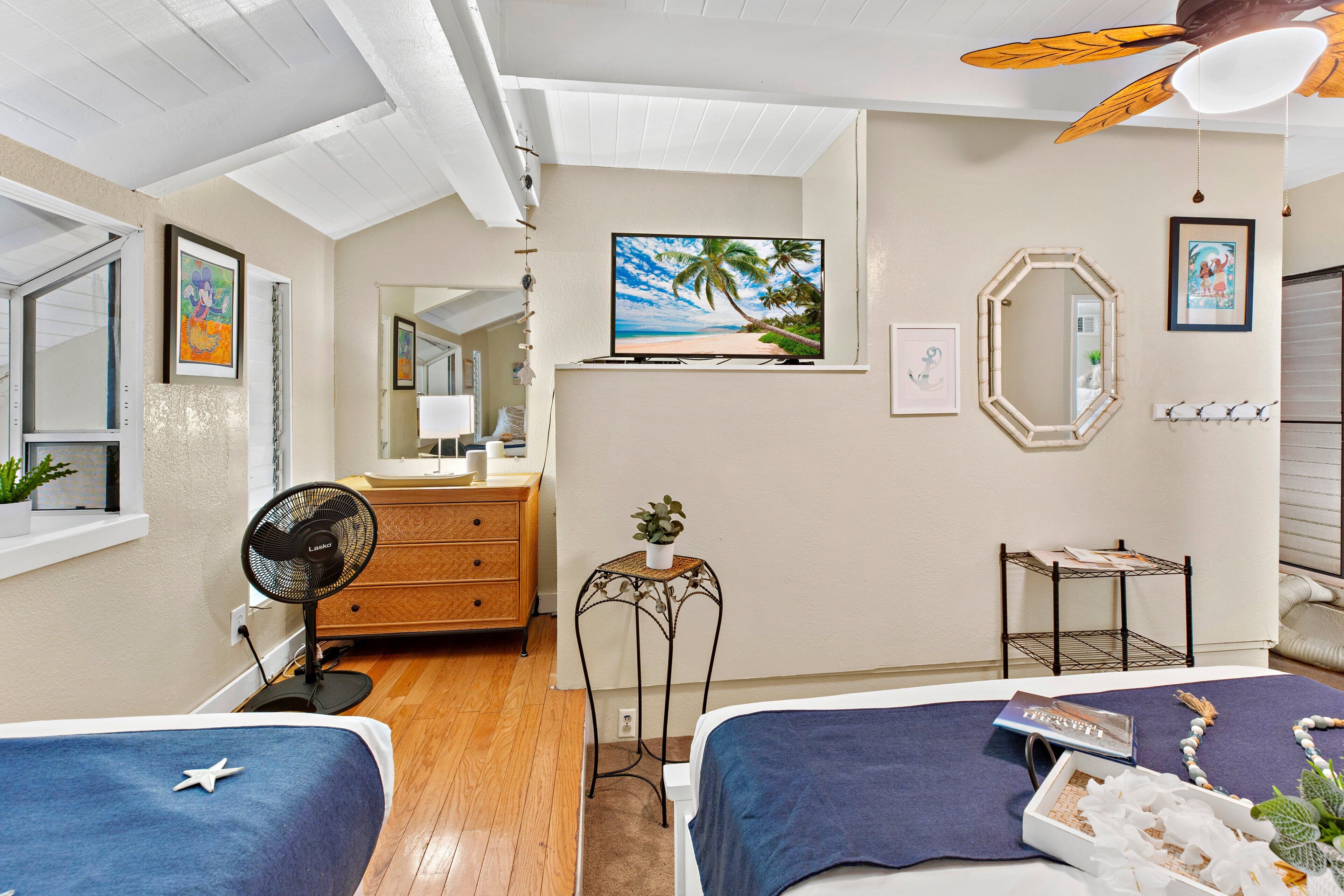 A bedroom with two beds, a ceiling fan, a TV, a small table with a plant, and a mirror on the wall.