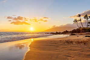 Une plage au coucher du soleil, avec des palmiers et des formations rocheuses.