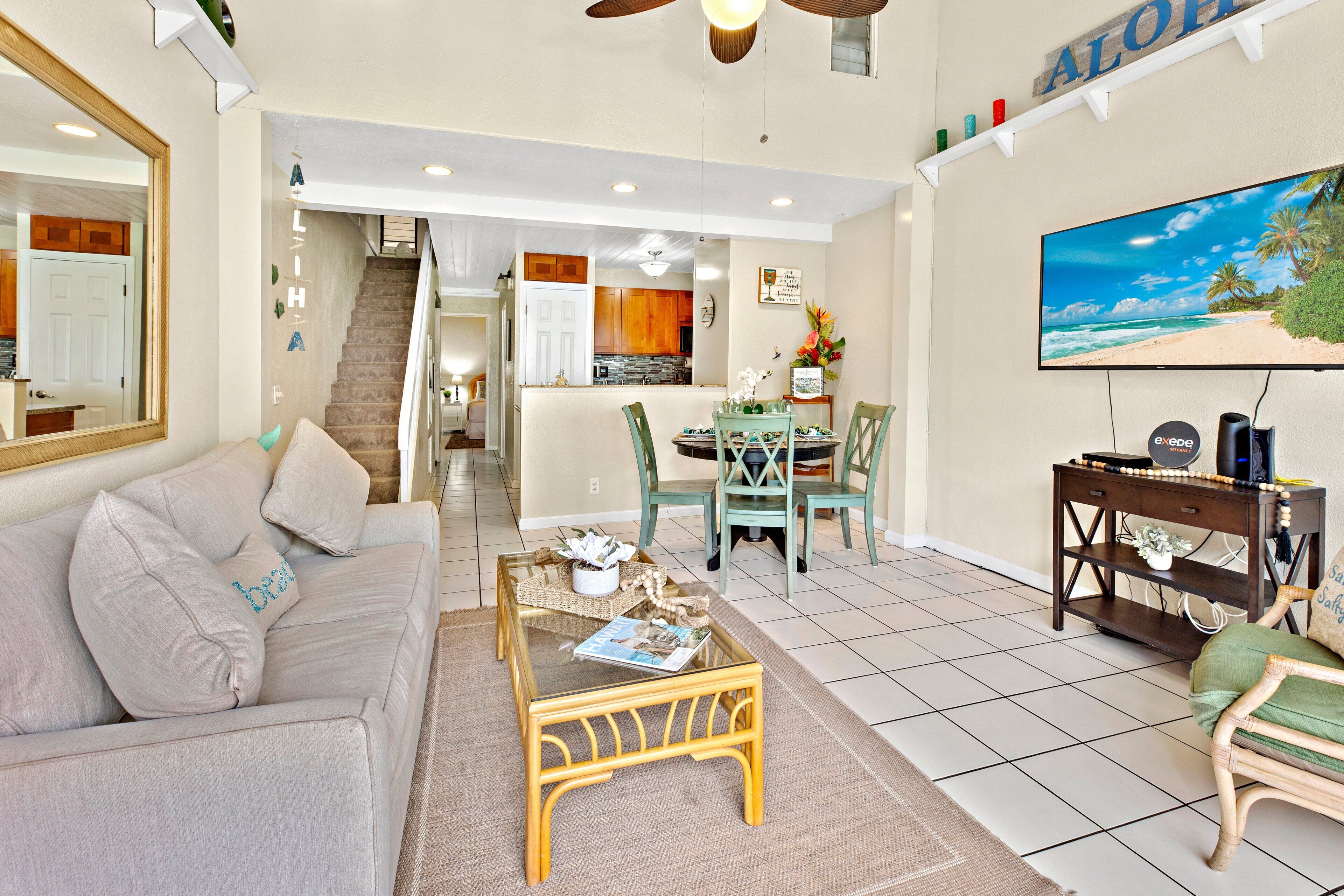 A living room with a sofa, coffee table, and TV. There's a staircase leading to a kitchen and dining area.