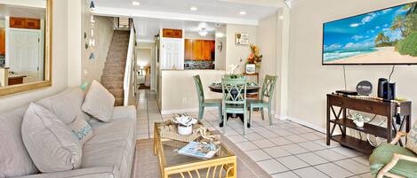 A living room with a sofa, coffee table, and TV. There's a staircase leading to a kitchen and dining area.