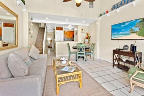 A living room with a sofa, coffee table, and TV. There's a staircase leading to a kitchen and dining area. - Top-Floor 2BR Tropical Escape Near the Beach (Kihei)