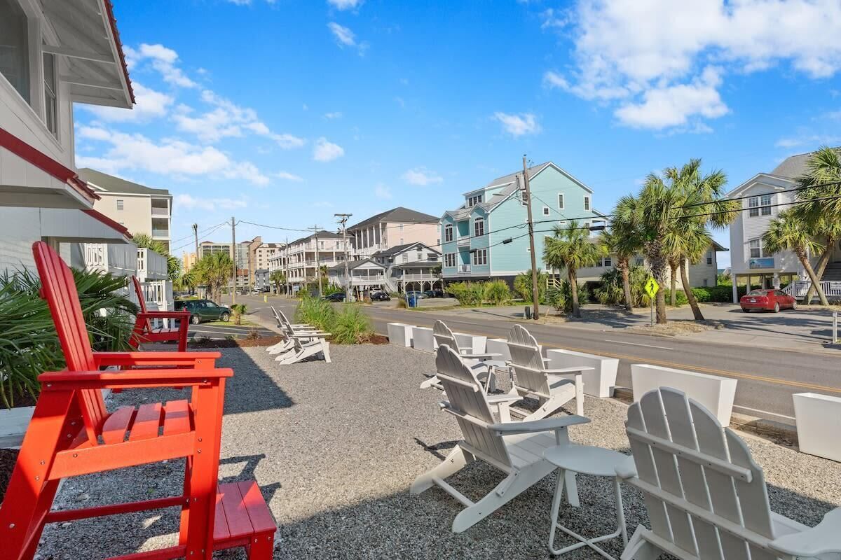 A beachfront property with a row of Adirondack chairs, a parking lot, and a street with cars and buildings in the background.