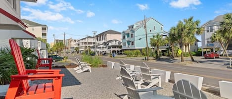 A beachfront property with a row of Adirondack chairs, a parking lot, and a street with cars and buildings in the background.