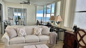 A spacious living room with a large sofa, ottomans, a coffee table, and a view of the ocean through large windows. - St. Nicole Condo in Pelican Bay, Naples, Florida with Gorgeous Gulf Views (Naples)