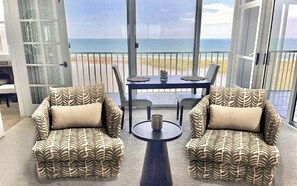 Een balkon met uitzicht op zee, twee gestreept fauteuils, een klein tafeltje en een eettafel met stoelen.