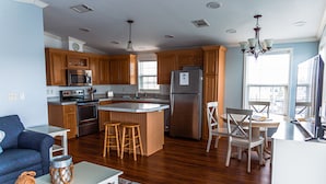 A modern kitchen with wooden cabinets, stainless steel appliances, a central island with stools, and a dining area with a chandelier. - Endless Water Views - Venture Out 526 (Cudjoe Key)