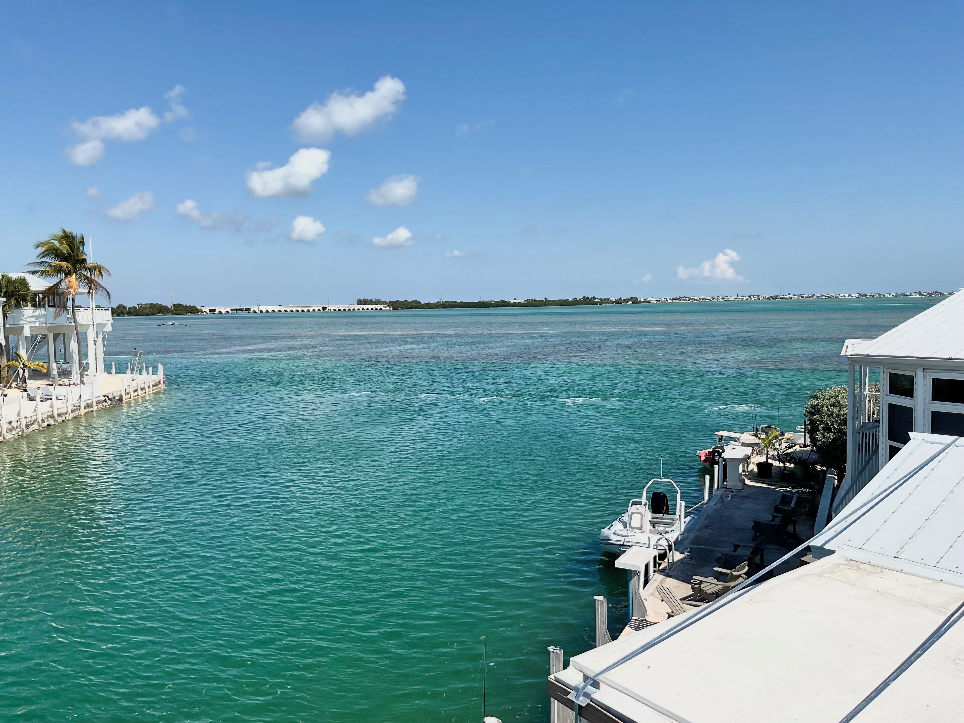 A marina with a docked boat, a white building with a balcony, and a clear view of the water and distant shoreline.