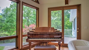 Un piano de cola en una habitación con ventanas amplias, una silla blanca y vistas a una terraza y árboles.
