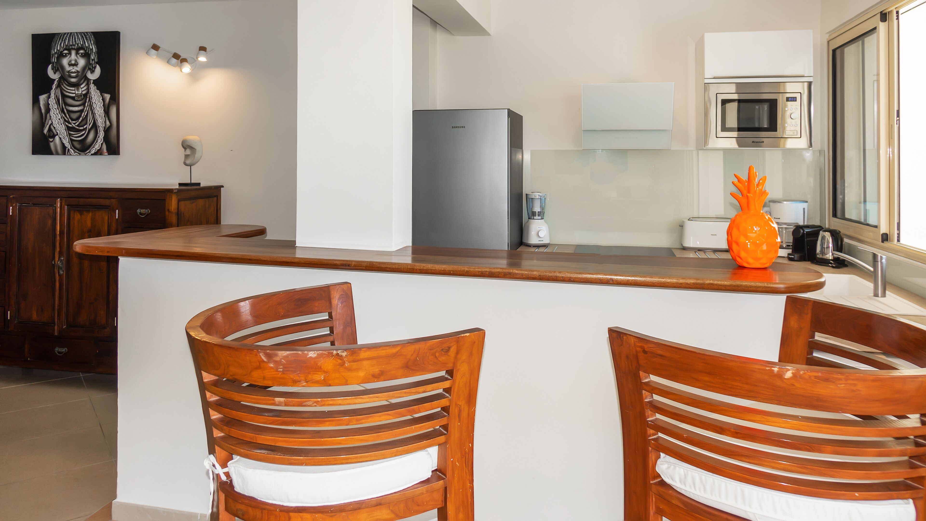 A kitchen with a wooden bar, two wooden chairs, a refrigerator, a microwave, and a blender.