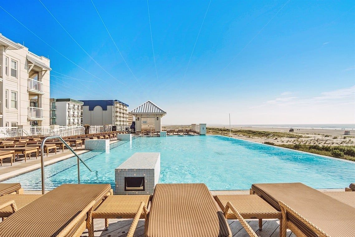 A swimming pool with lounge chairs, a building with balconies, and a beach in the background.