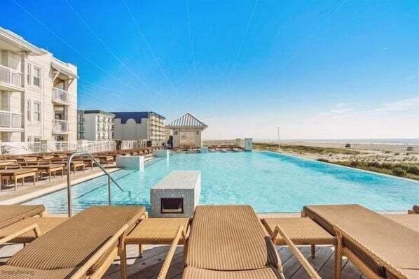 Uma área de piscina com espreguiçadeiras, um edifício com varandas e um céu claro.