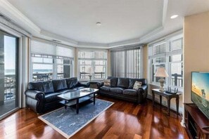 Uma sala de estar com um sofá de couro preto, uma mesa de centro e uma TV sobre um suporte.