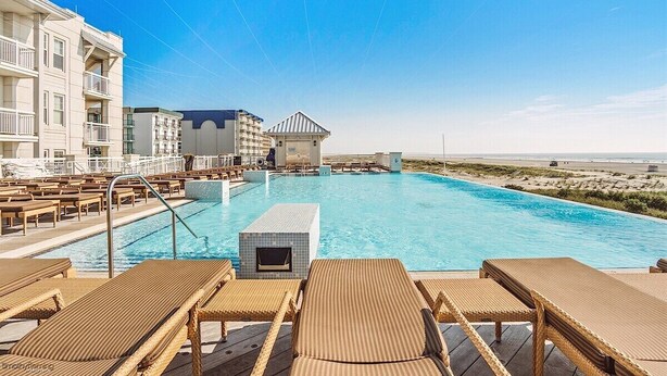Zona de piscina con sillones, un edificio con balcones y vista a la playa.