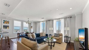 Una sala de estar moderna con sofá, sillones, una mesa de centro y televisor. Hay ventanas amplias con cortinas, un comedor con mesa de vidrio y vistas a la ciudad.