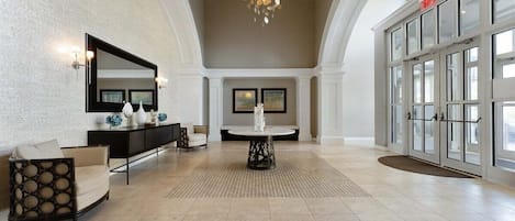 Een ruime lobby met een hoge plafond, een kroonluchter, een receptiebalie, een zithoek en een grote glazen deur als ingang.