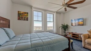 A bedroom with a bed, a ceiling fan, two windows, a painting, a television, and a plant.