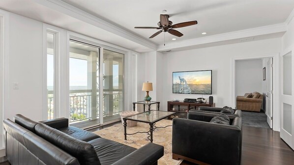A spacious living room with a large glass door leading to a balcony, a ceiling fan, and a flat-screen TV mounted on the wall.