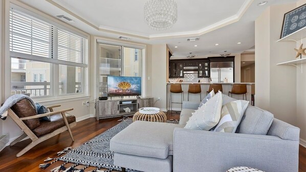 A modern living room with a sofa, armchair, and ottoman, a dining area with bar stools, and a kitchen with a large island.