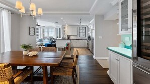 A modern kitchen with white cabinets, a stainless steel refrigerator, and a dining area with a wooden table and wicker chairs. - Residence 102 at The Grand (Wildwood)
