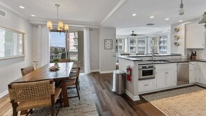 A spacious kitchen with a large island, white cabinetry, and a dining area with a wooden table and wicker chairs. - Residence 102 at The Grand (Wildwood)