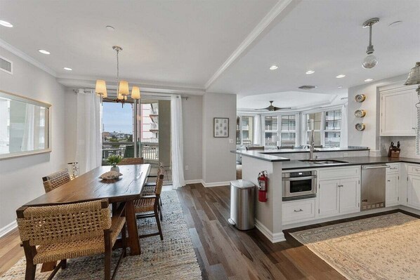 A spacious kitchen with a large island, white cabinetry, and a dining area with a wooden table and wicker chairs. - Spacious 3BR Residence Near Sundeck Amenities (Wildwood)