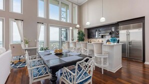 Un vaste espace de salle à manger avec une grande table et des chaises blanches, une cuisine équipée d’appareils modernes et de grandes fenêtres offrant une vue dégagée.