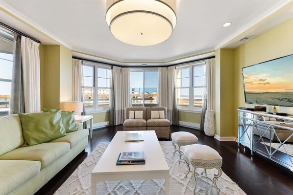 Una sala de estar moderna con sofá, mesa de centro y sillones, ventanas amplias con cortinas, televisor y vista al mar.