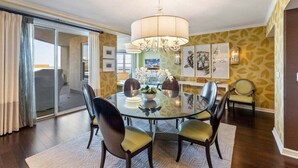 Uma sala de jantar com uma mesa redonda de vidro, quatro cadeiras, um candelabro e vista através de uma porta de vidro.