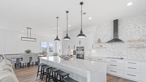 A modern kitchen with a large island, pendant lights, and white cabinetry. - Carolina Blue Skies is 2nd Row, Steps to the Beach, Ocean Views and New Construction (Holden Beach)
